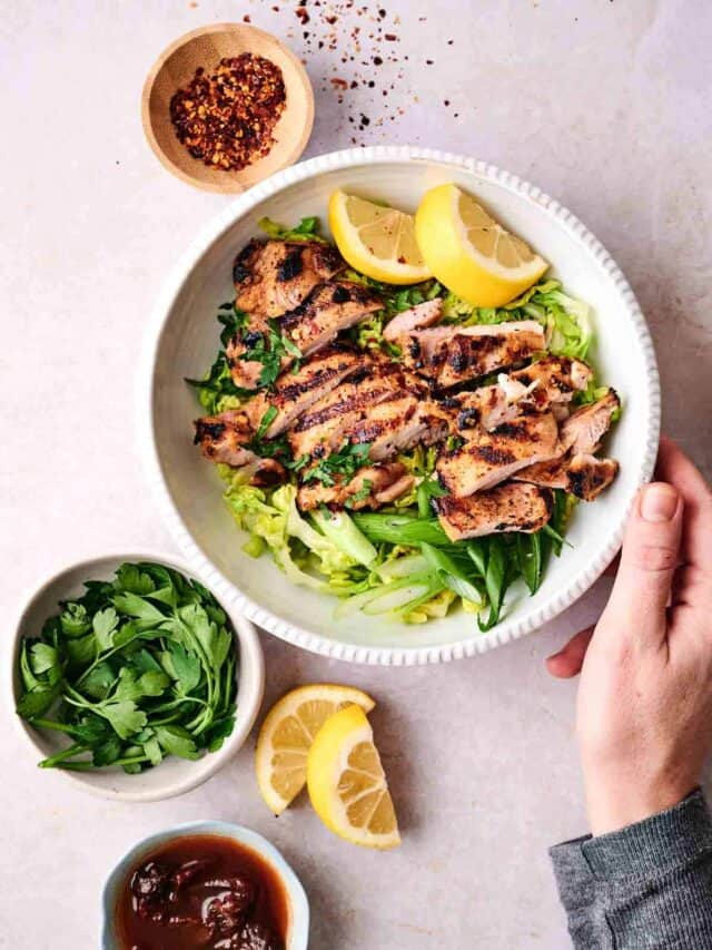 A person holding a plate of grilled chicken slices on a bed of greens garnished with lemon wedges, with bowls of herbs and sauce nearby.