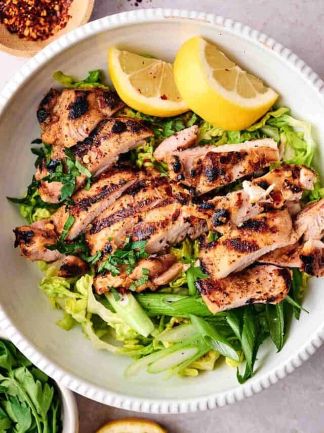 A bowl of grilled chicken slices on a bed of greens, topped with lemon wedges and herbs.