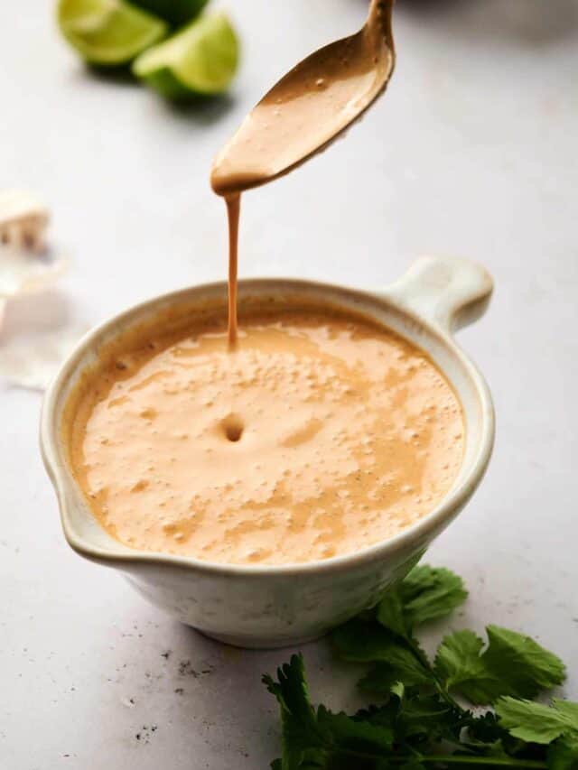 A creamy dressing being drizzled from a spoon into a small bowl, with citrus and herbs nearby.