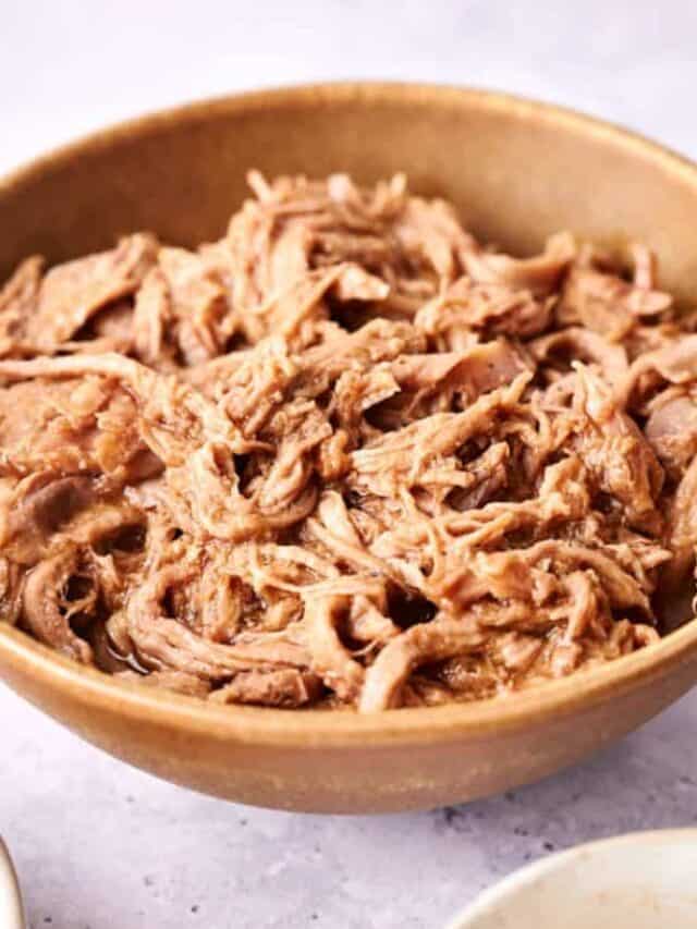 A bowl of shredded, cooked pork in a light-colored ceramic dish on a marble surface.