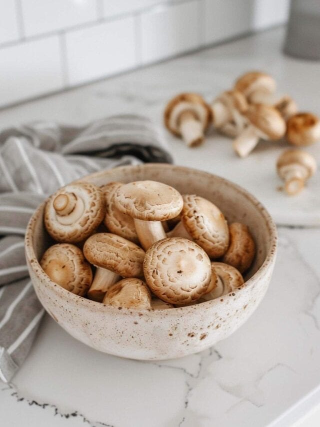 A bowl of raw mushrooms on a kitchen countertop, with more mushrooms scattered around it.