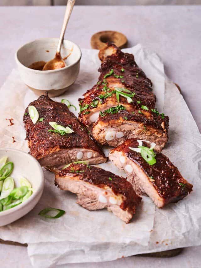 A serving of sliced, glazed ribs garnished with green onions, accompanied by a small bowl of sauce and another bowl of sliced green onions on a parchment-lined surface.