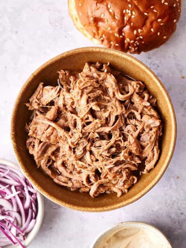 A bowl of shredded pork sits on a table alongside a sesame seed bun, a dish of sliced red onions, and a container of sauce.