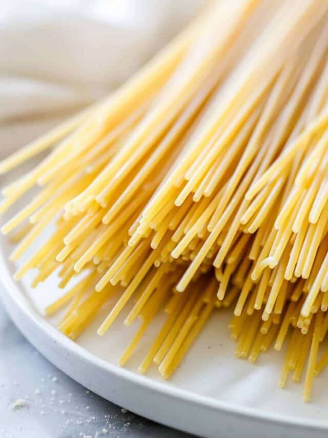Close-up image of uncooked spaghetti noodles piled on a white plate, with some strands extending over the edge.