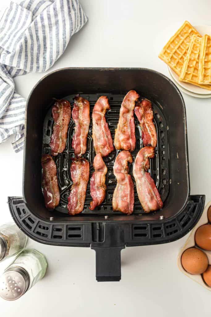Slices of Bacon strips cooking in an air fryer, surrounded by eggs, a salt shaker, and waffles on a white surface.