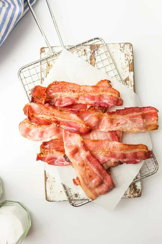 Air Fryer bacon strips draining on a wire rack over a white cutting board, with a blue-striped towel on the side.