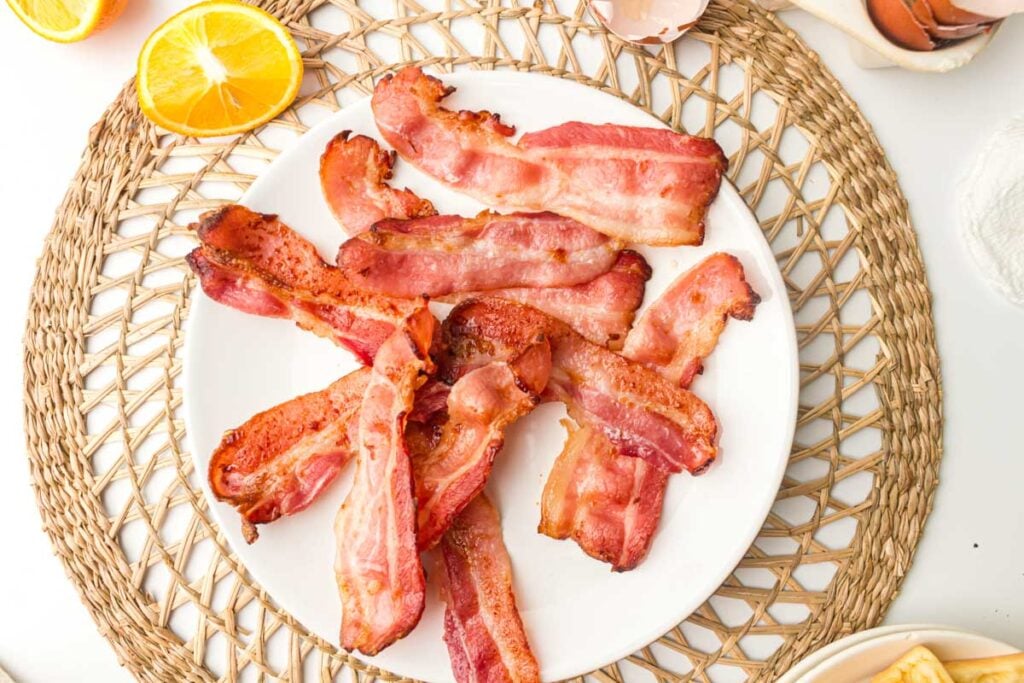 A white plate with several crispy Air Fryer Bacon strips on a woven placemat, with orange slices and eggshells nearby.