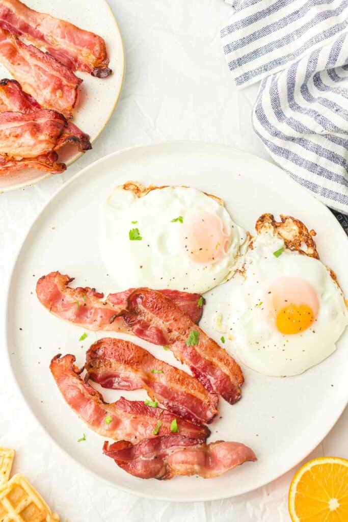 Two sunny-side-up eggs and air fryer bacon strips on a white plate, accompanied by a side plate of bacon and a striped napkin.