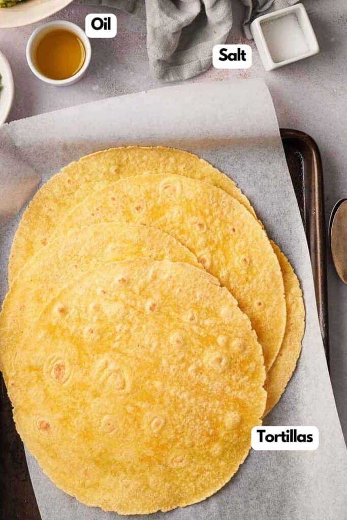 Three corn tortillas stacked on parchment paper, with labeled ingredients including oil and salt nearby on a kitchen countertop.