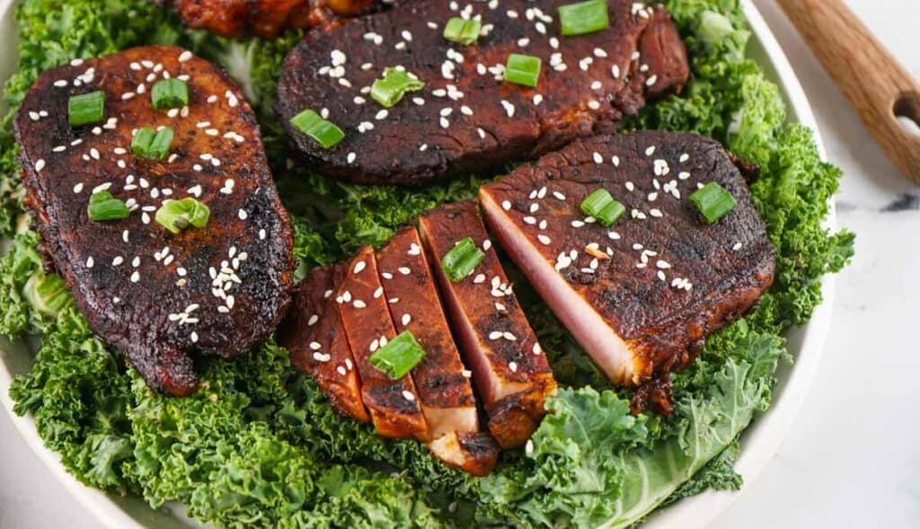 Grilled asian style pork chops with barbecue sauce garnished with sesame seeds and green onions, served on a bed of kale.