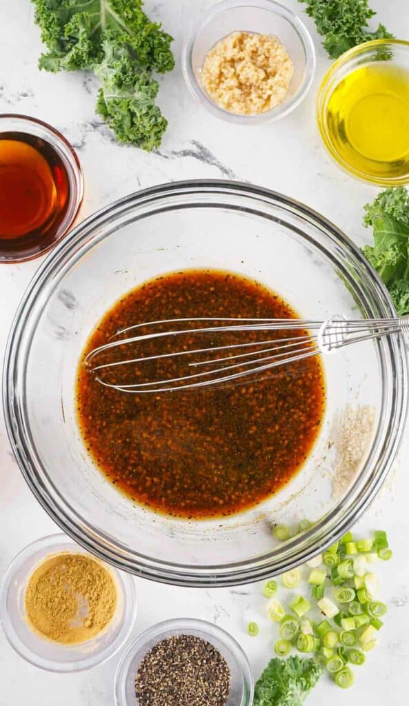 A clear glass bowl containing a marinade for Asian style pork chops with a whisk on a white surface surrounded by ingredients like kale, oil, and spices.