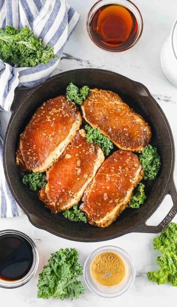 Asian-style glazed pork chops in a skillet, surrounded by kale, with sauces and spices on the side.