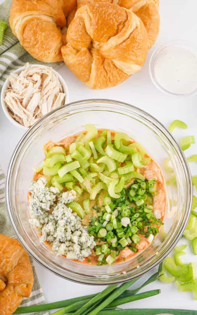 A bowl of buffalo chicken salad topped with celery, green onions, and blue cheese, surrounded by croissants and fresh celery sticks.