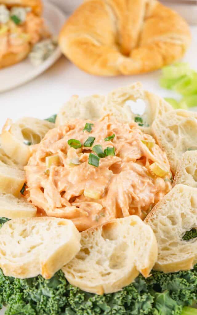 Buffalo chicken salad served with sliced baguette and celery on a white plate, garnished with chopped green onions.