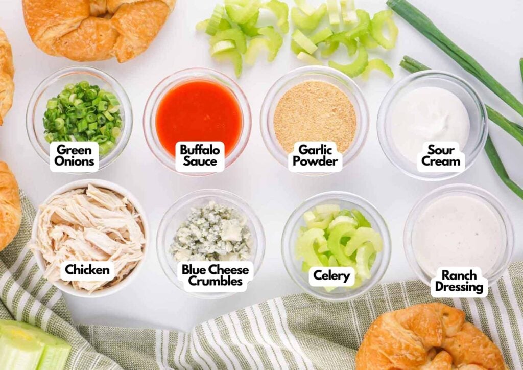 Top view of labeled ingredients in bowls for Buffalo Chicken Salad: green onions, buffalo sauce, garlic powder, sour cream, chicken, blue cheese crumbles, celery, and ranch dressing, with croissants on the side.