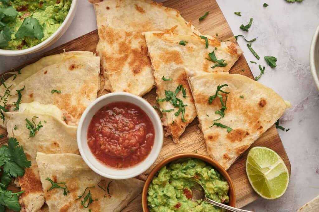 Sliced quesadillas on a wooden board, served with bowls of guacamole and salsa, garnished with cilantro and lime wedges.