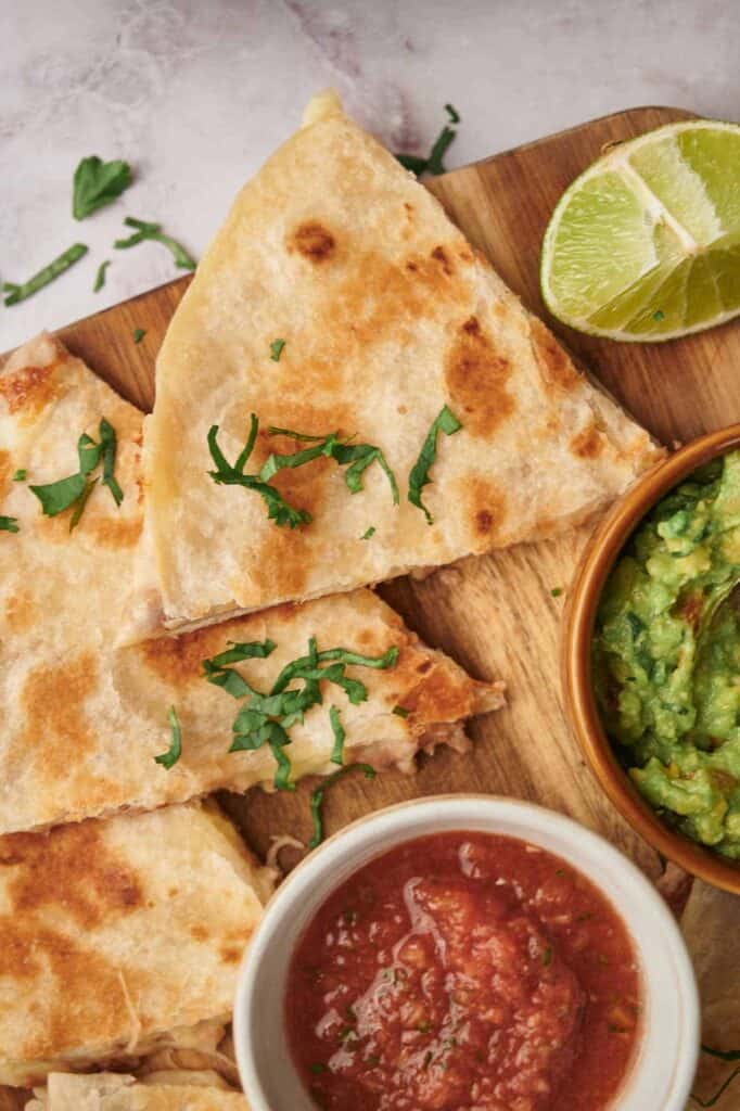 A quesadilla sliced into triangles, garnished with cilantro, served with bowls of guacamole and salsa, with a lime wedge on the side.