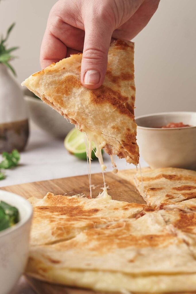 A hand lifts a gooey cheese quesadilla slice from a wooden board, with melted cheese stretching; bowls of salsa and fresh greens are nearby.