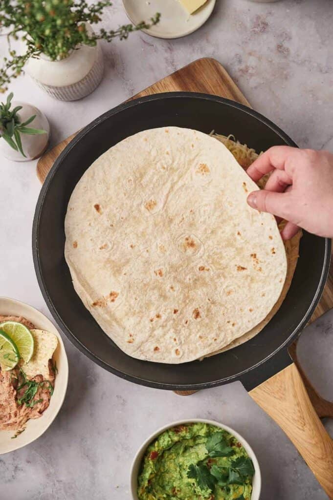 Hand placing a large tortilla on a skillet, with bowls of guacamole and lime aside, seen from an overhead perspective.
