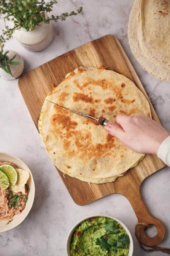 A person slicing a stack of quesadillas on a wooden cutting board, with tortillas and guacamole nearby.