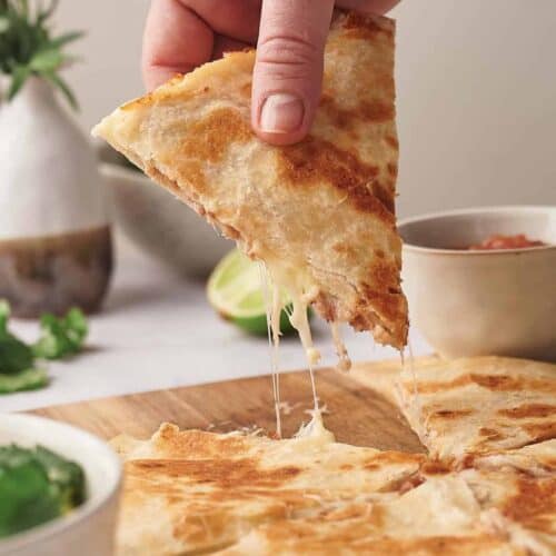 A hand lifting a slice of cheesy quesadilla, with melted cheese stretching between the slice and the main portion on a plate, accompanied by dipping sauces and lime.