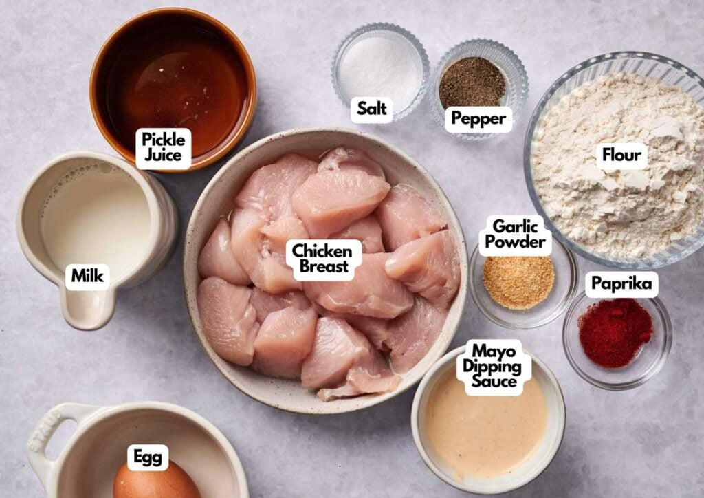 Overhead view of labeled ingredients in bowls and cups for making Chick-fil-A chicken nuggets, including chicken breast, pickle juice, milk, egg, mayo dipping sauce, salt, pepper, flour, garlic powder, and paprika.