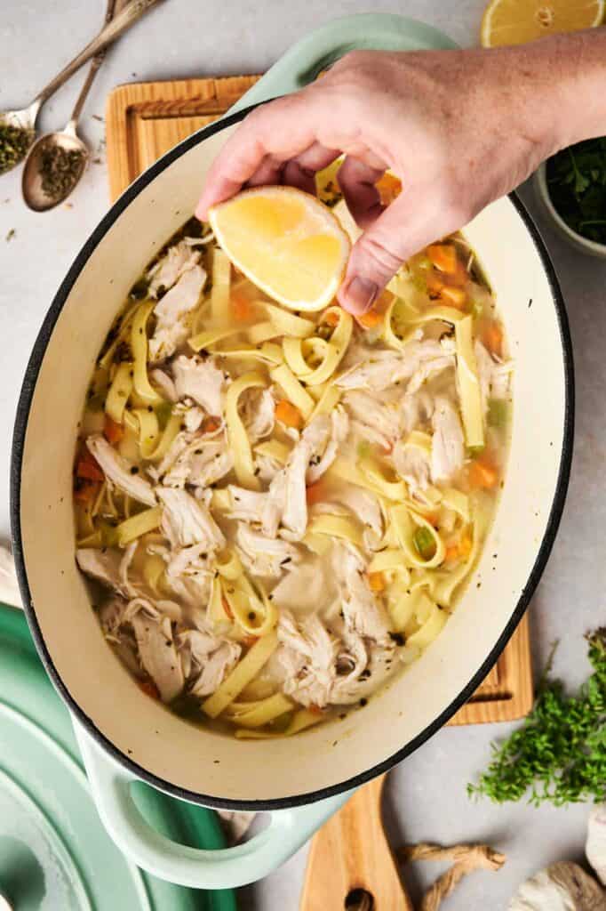 A hand adds a burst of flavor by squeezing a lemon slice over a pot of savory chicken noodle soup, rich with shredded chicken, vibrant vegetables, and perfectly tender noodles.