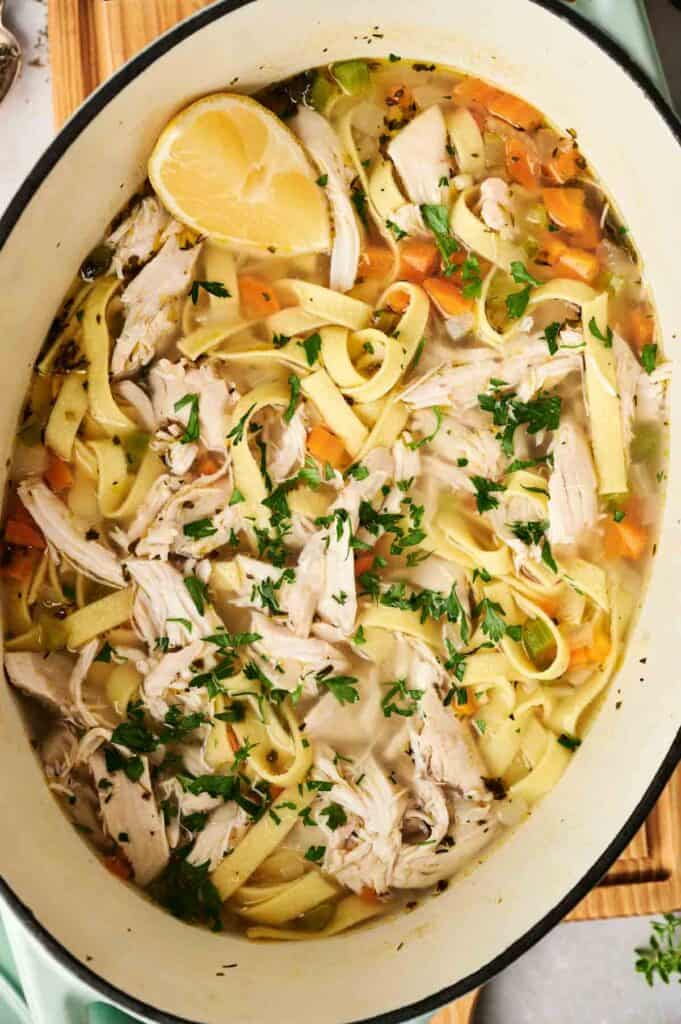 A steaming pot of chicken noodle soup brimming with shredded chicken, delicate noodles, vibrant carrots, and crisp celery is topped with fresh herbs and garnished with a zesty lemon wedge.