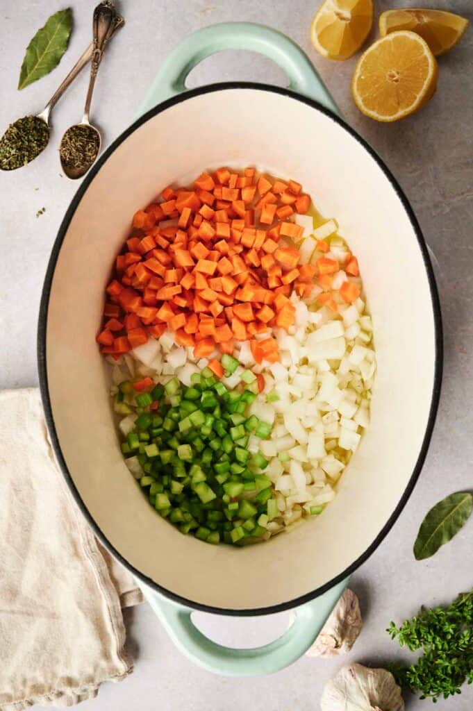 Chopped carrots, celery, and onions simmer in a large pot of chicken noodle soup. Around the pot are herbs, two lemon halves, spices, and garlic cloves on a light surface.