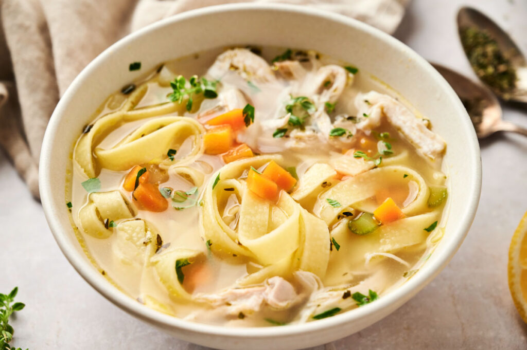 This comforting bowl of chicken noodle soup features wide noodles, diced carrots, celery, and fresh herbs in a light broth, all served on a table with a spoon nearby.