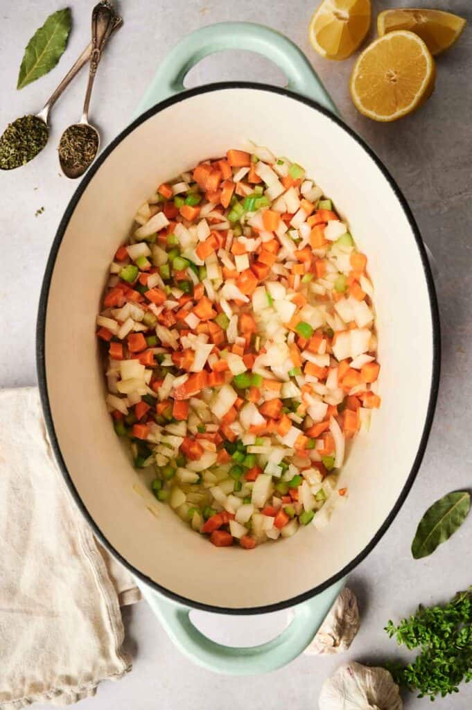 Chopped onions, carrots, and celery sit in a pot on a kitchen counter, ready to transform into comforting chicken noodle soup. Nearby are lemon halves, herbs, a cloth, and garlic.