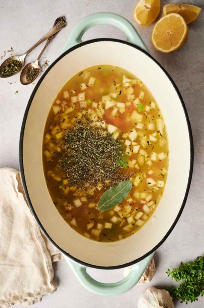 A pot of chicken noodle soup with diced vegetables, a bay leaf, and herbs on top. Lemons, garlic, and spoons with spices are nearby on a gray surface.
