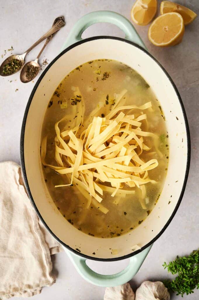 A pot of chicken noodle soup with wide noodles in broth, surrounded by lemon halves, herbs, garlic, and a cloth napkin on a light surface.