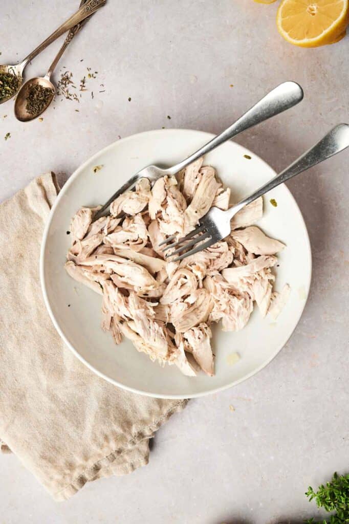 A bowl of shredded chicken with two forks rests on a light countertop, reminiscent of the flavors found in a comforting chicken noodle soup. It's surrounded by fresh herbs, a lemon half, and a beige napkin, creating an inviting scene that promises warmth and deliciousness.