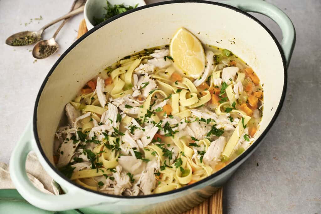 A savory pot of chicken noodle soup, featuring tender shredded chicken, vibrant carrots, and a sprinkle of parsley, all beautifully garnished with a lemon wedge.