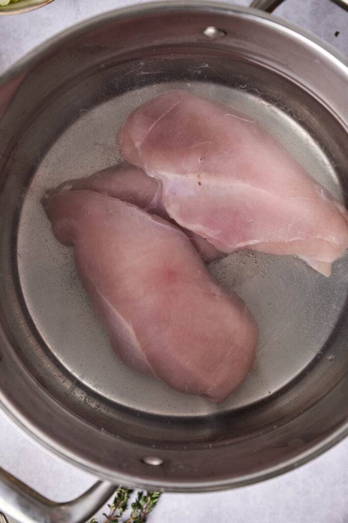 Two raw chicken breasts soaking in brine in a stainless steel bowl, prepped for chicken pot pie.