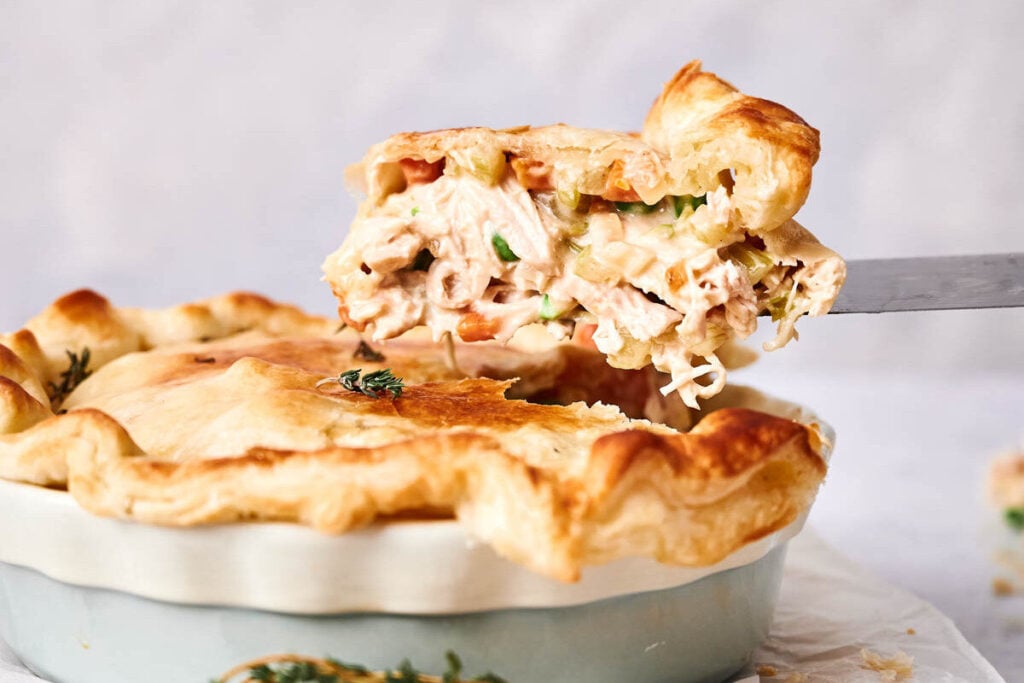A slice of Chicken Pot Pie with a golden, flaky crust is being lifted from a pie dish, revealing its creamy filling packed with chicken and vegetables.