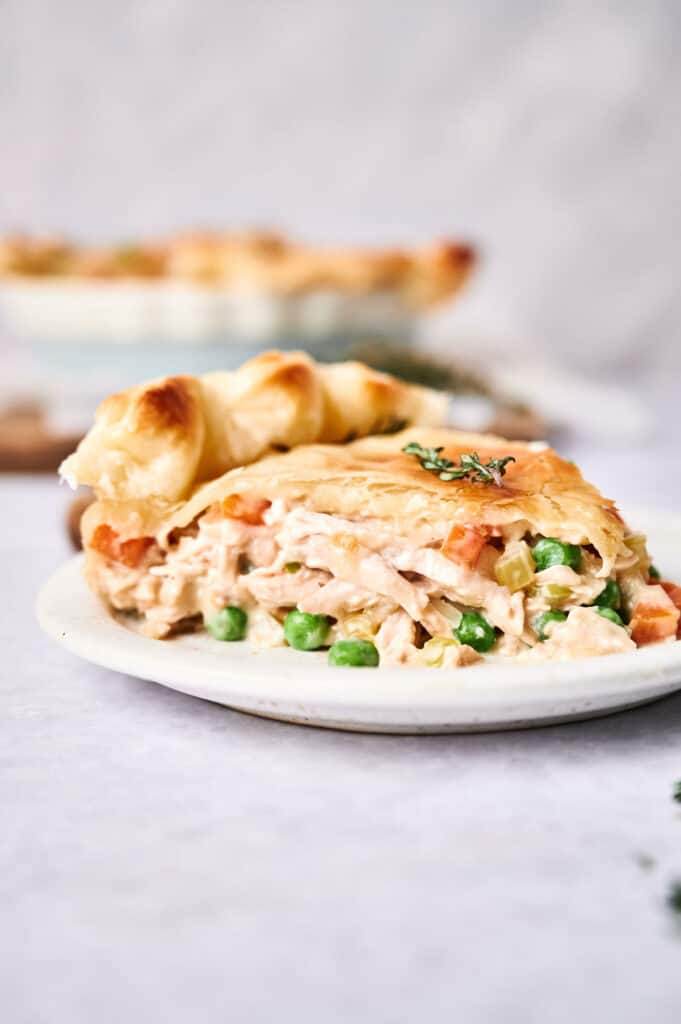 A plate with a slice of chicken pot pie, displaying its creamy filling with vegetables, in front of a chicken pot pie dish.
