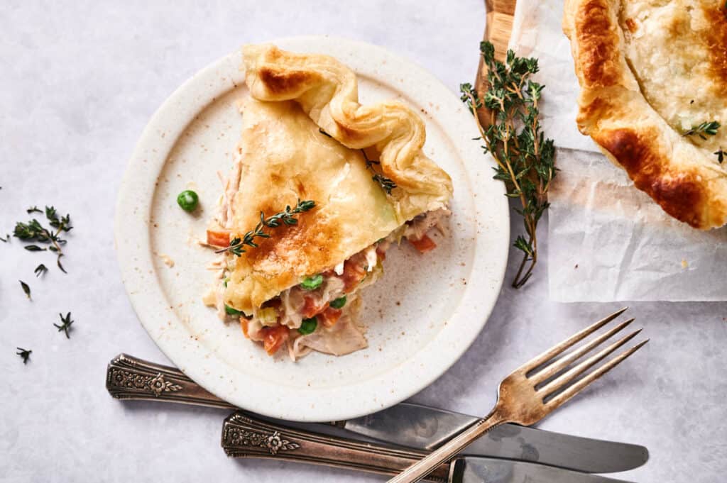 A slice of chicken pot pie served on a white plate with fresh herbs and cutlery on the side.