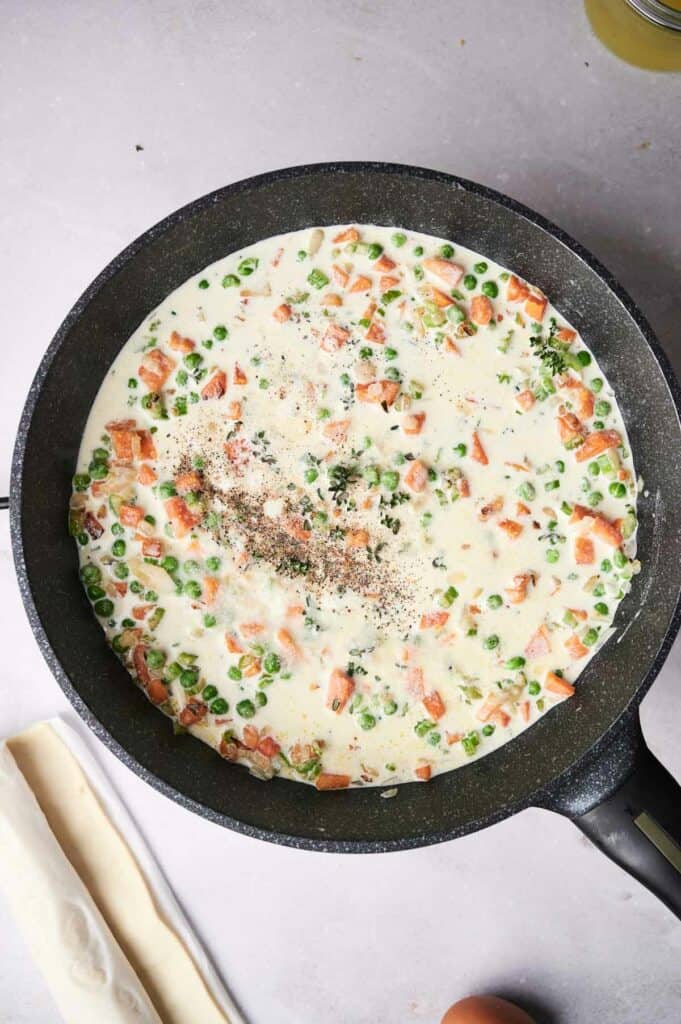 A skillet containing creamy sauce with carrots, peas, and spices, on a light-colored surface with breadsticks beside it.