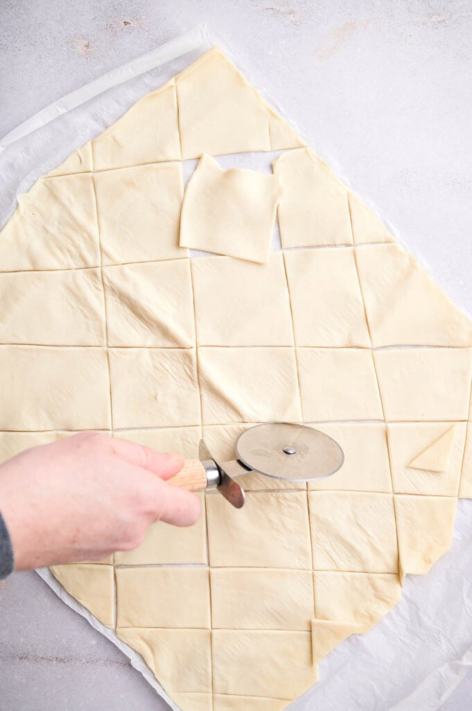 A person using a pizza cutter to slice a sheet of dough into small triangles on a kitchen surface.