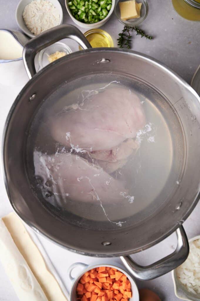 Raw chicken breasts submerged in boiling water in a pot, surrounded by ingredients like carrots, leeks, and herbs on a kitchen counter.