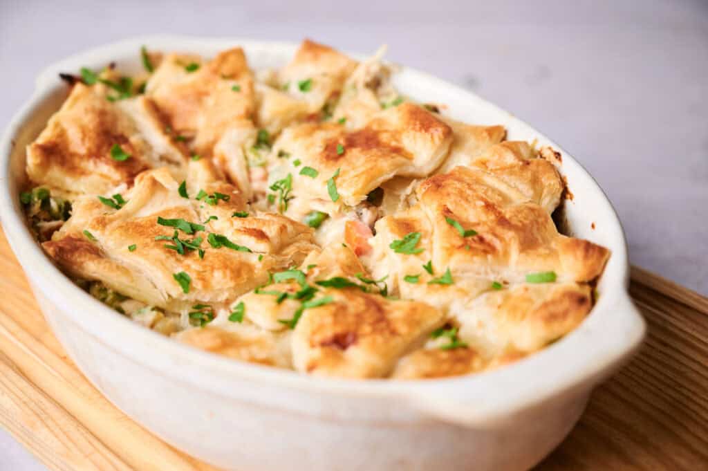 A chicken pot pie in a white ceramic dish, topped with golden-brown pastry and garnished with chopped parsley.