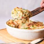 A hand lifting a serving of creamy, chicken pot pie casserole from a white oval baking dish, garnished with parsley.