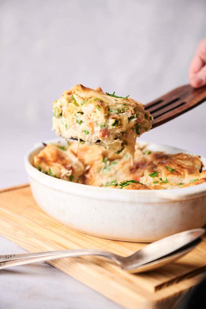 A hand lifting a serving of creamy, vegetable-filled casserole from a white oval baking dish, garnished with parsley.