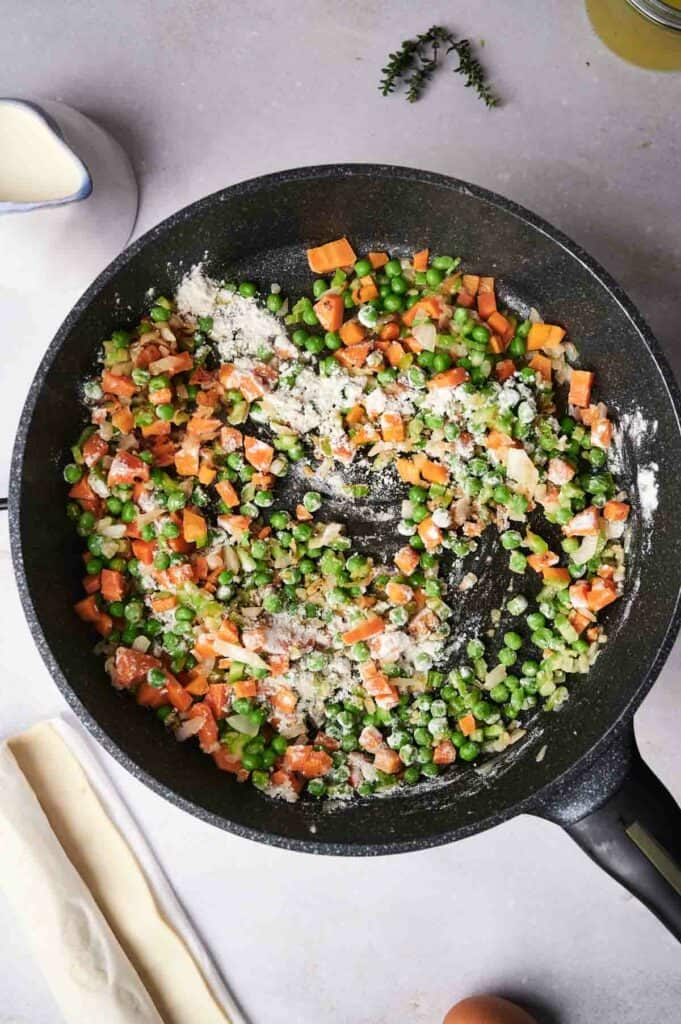 A skillet containing mixed diced vegetables, including carrots, green peas, and onions, sautéing on a stove with seasonings visible.