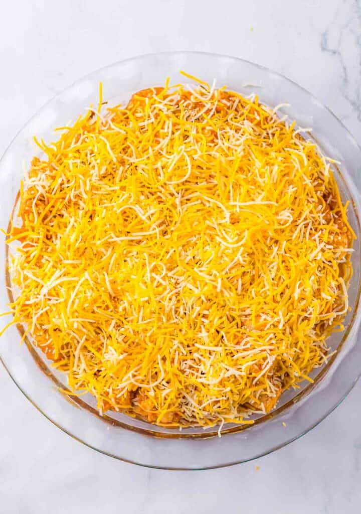 A glass pie dish filled with a thick layer of shredded yellow and white cheese on top of a chicken tamale filling, placed on a white marble surface.