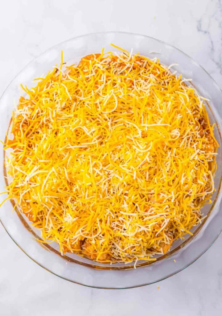 A glass pie dish filled with a thick layer of shredded yellow and white cheese on top of a chicken tamale filling, placed on a white marble surface.