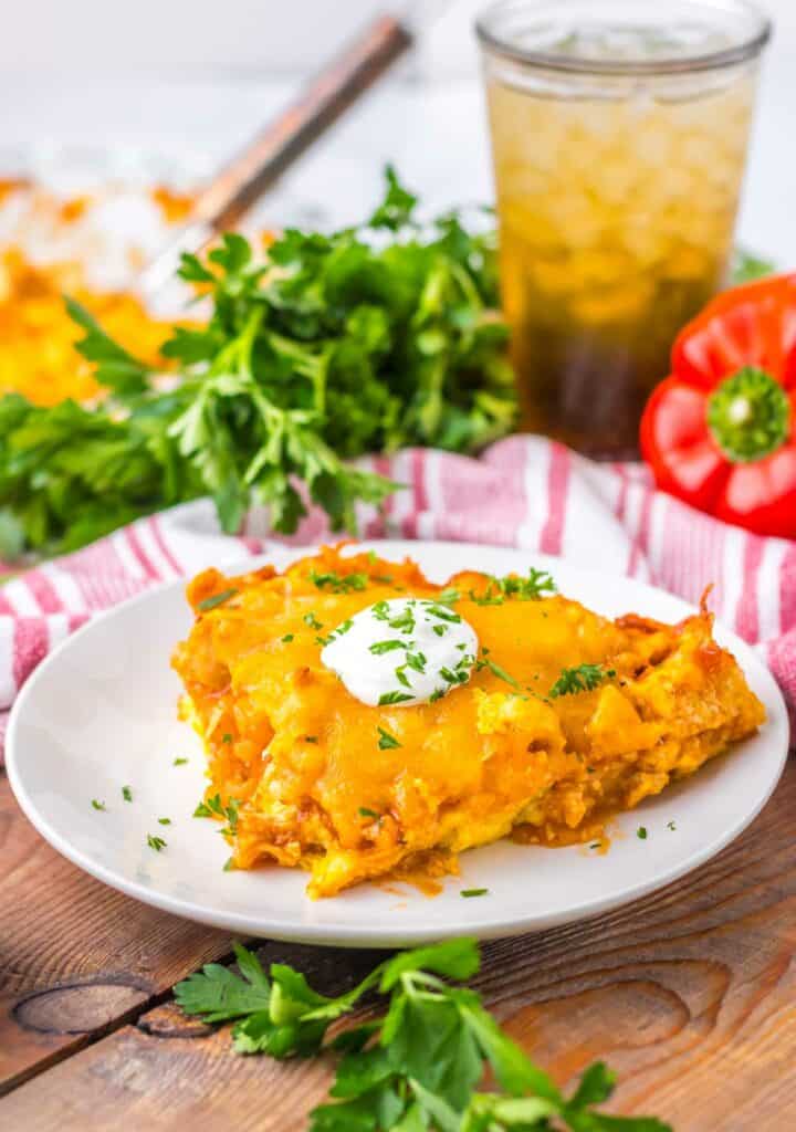 A slice of cheesy chicken tamale pie topped with sour cream on a plate, garnished with parsley, with a drink and fresh vegetables in the background.
