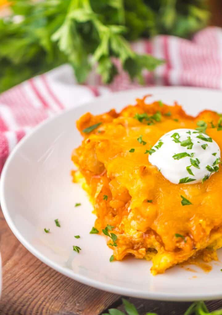 A slice of cheesy chicken tamale pie on a white plate garnished with a dollop of sour cream and chopped herbs, with a striped napkin on the side.